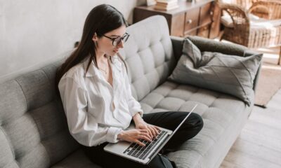 A woman with glasses sitting cross legged on a gray couch working on laptop
