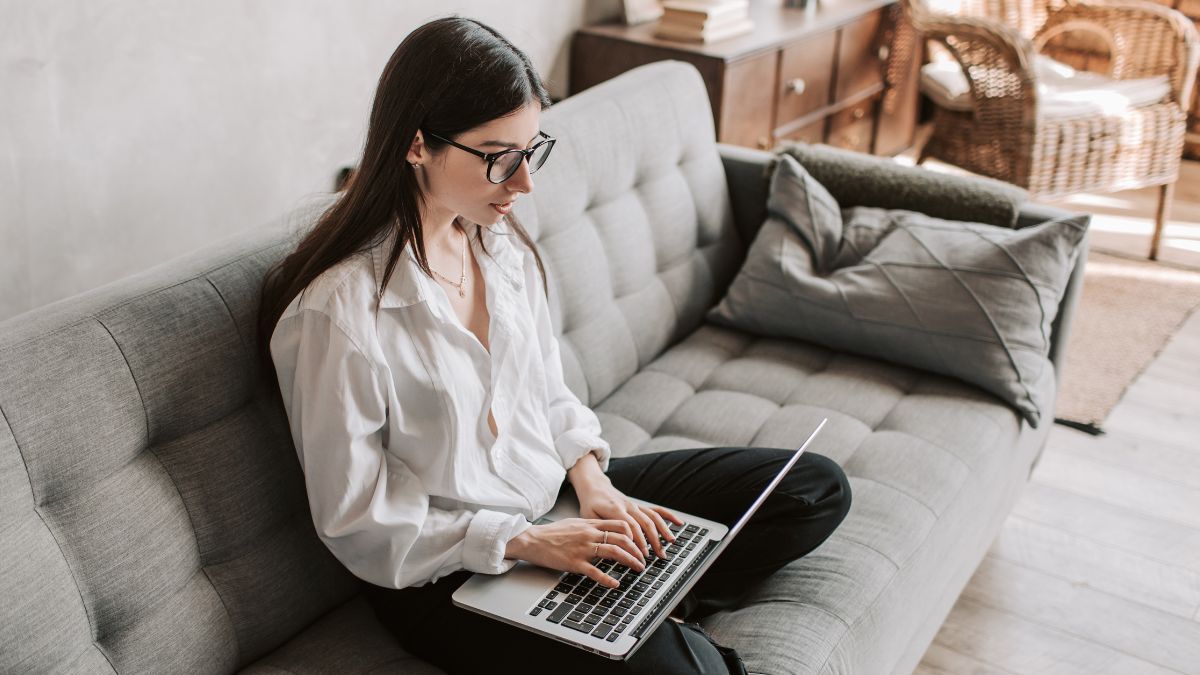 A woman with glasses sitting cross legged on a gray couch working on laptop